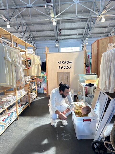 Client kneeling next to his toddler surrounded by displays and hanging t-shirts made by the business.